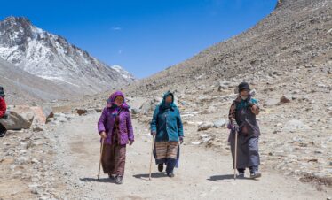 Three women trek through Ngari's mountainous terrain, clad in traditional attire and using walking sticks.